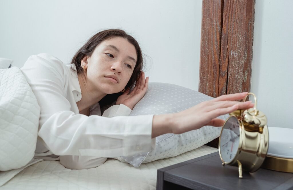 A tired person sitting on the edge of a bed in the early morning, illustrating the challenge of waking before feeling fully rested.