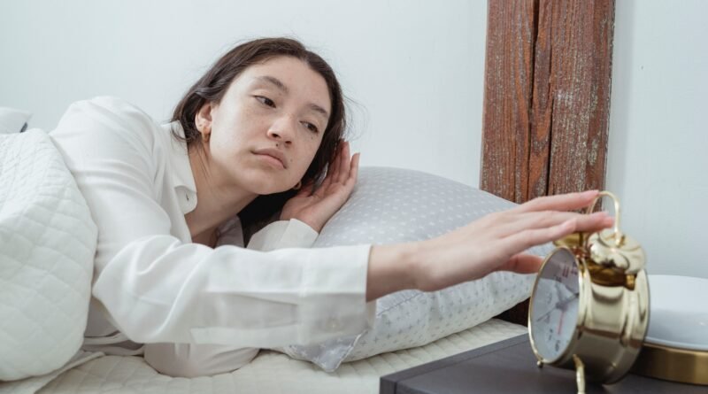 A tired person sitting on the edge of a bed in the early morning, illustrating the challenge of waking before feeling fully rested.